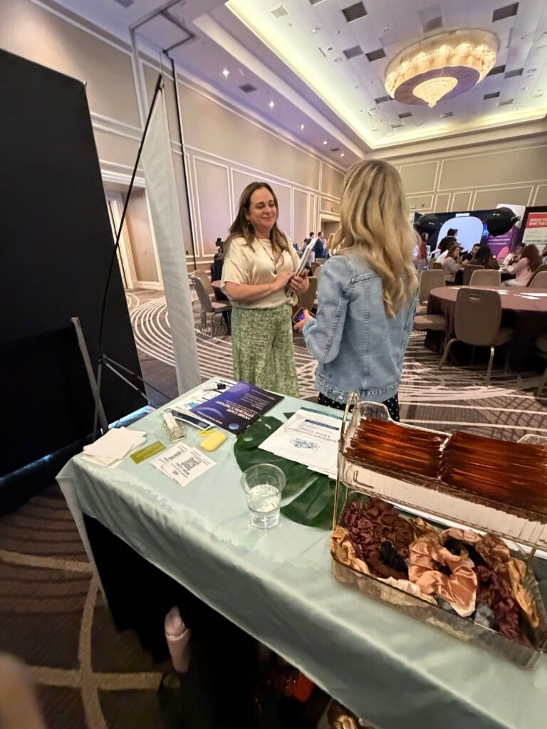 Two women having a conversation at a conference exhibition booth with brochures and informational materials on the table.