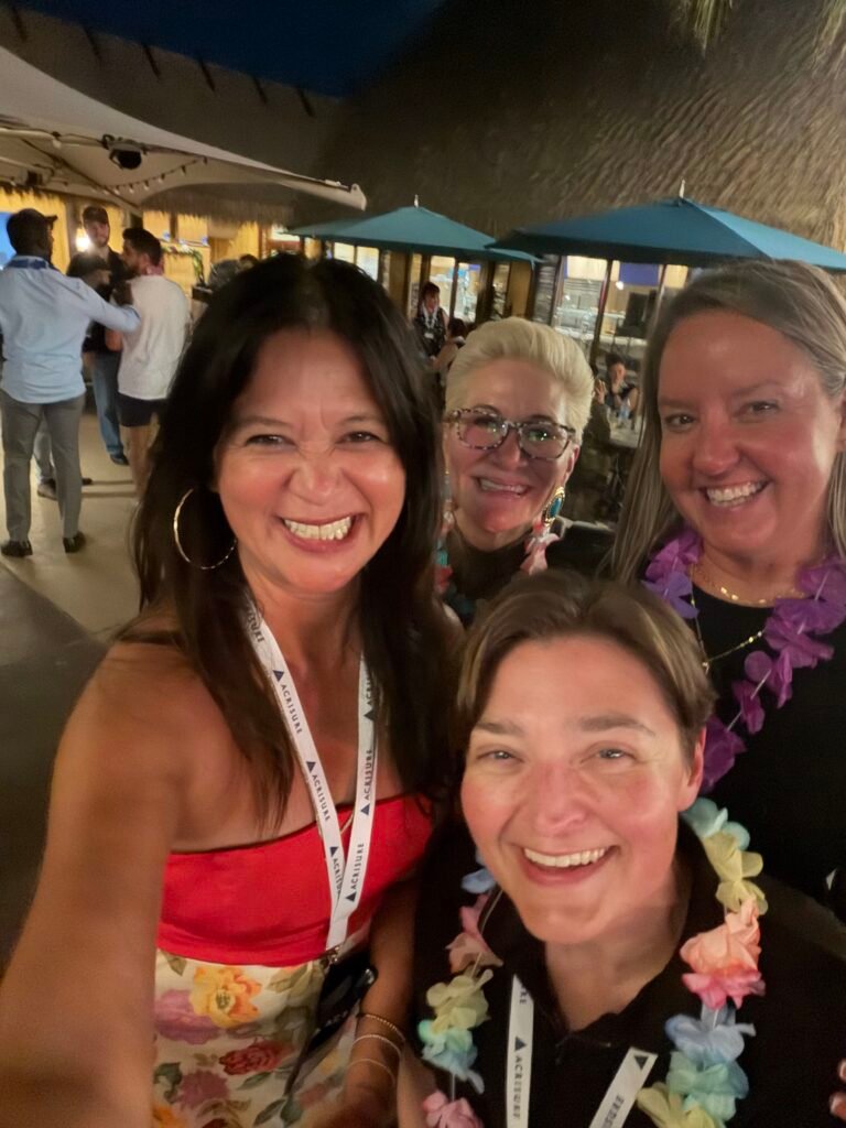 Four women smiling for a group selfie while wearing colorful leis at a tropical-themed evening networking event in Orlando.