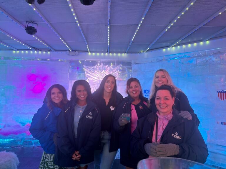 Six women wearing insulated jackets pose inside an ice bar illuminated by colorful blue and purple lights.