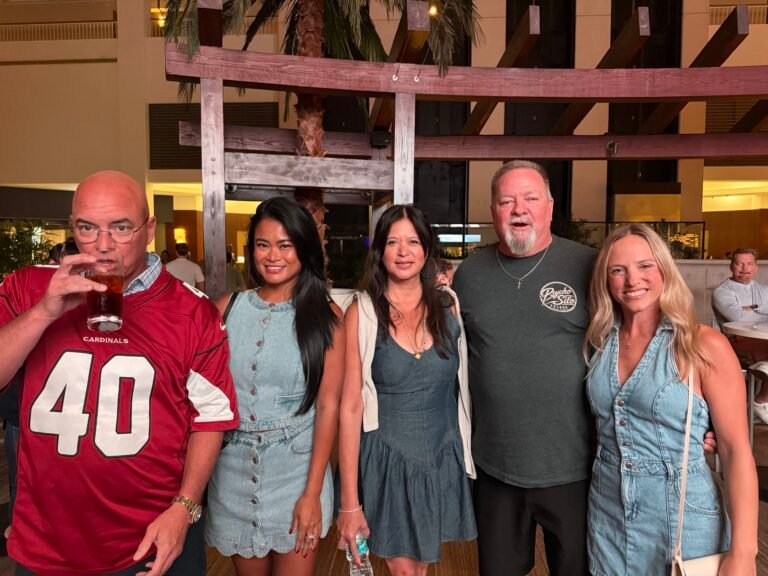 Five adults pose together in a hotel atrium during an evening social gathering, with warm lighting and wooden architectural details.