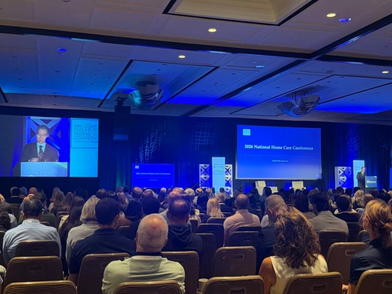 Large audience attending the HCAOA 2026 National Home Care Conference in a ballroom with blue stage lighting and presentation screens.
